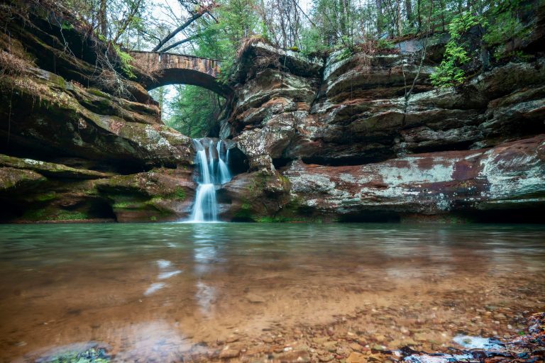 A serene view of Upper Falls at Old Man's Cave Trail in Hocking Hills State Park, Ohio, featuring a smooth waterfall flowing into a shallow, clear green pool, framed by ancient rock walls and an arched stone bridge overhead, surrounded by dense forest.