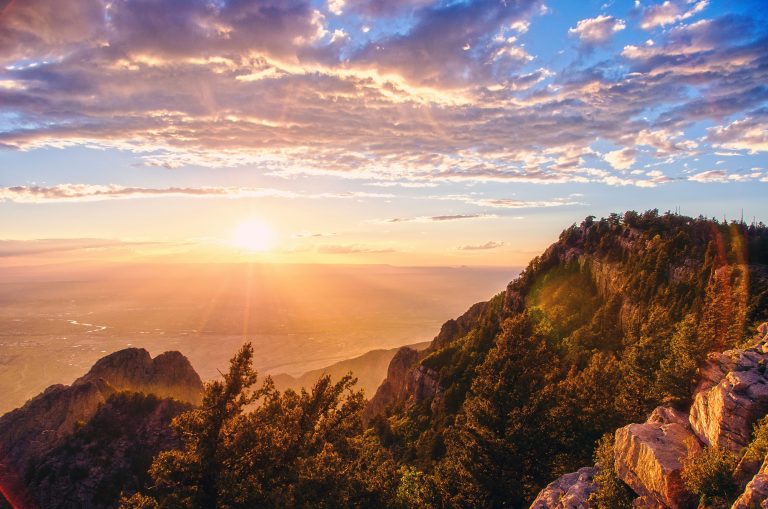 A dramatic high-altitude desert sunset scene in New Mexico's Sandia Mountains, an area where the Pino Trail is located. The view overlooks a sprawling valley, bathed in the warm glow of the setting sun