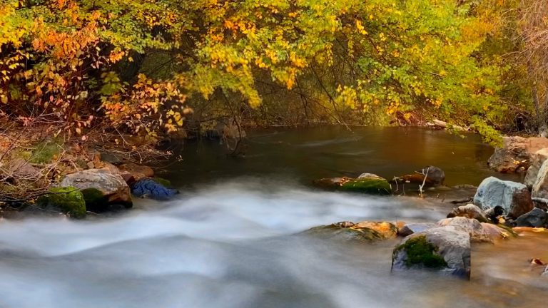 A long exposure shot of a flowing stream with a hazy, smooth appearance, surrounded by rocks and vibrant fall foliage in shades of gold and orange, likely on the Silver Lake Loop Trail in Utah.