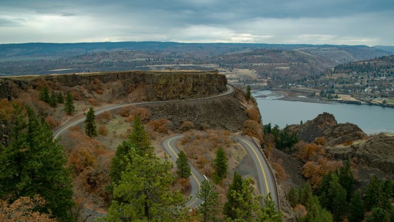 The iconic winding road at Rowena Crest Viewpoint, also known as part of the Tom McCall Point trail area, overlooking the Columbia River Gorge with distant hills under a dramatic cloudy sky in Oregon